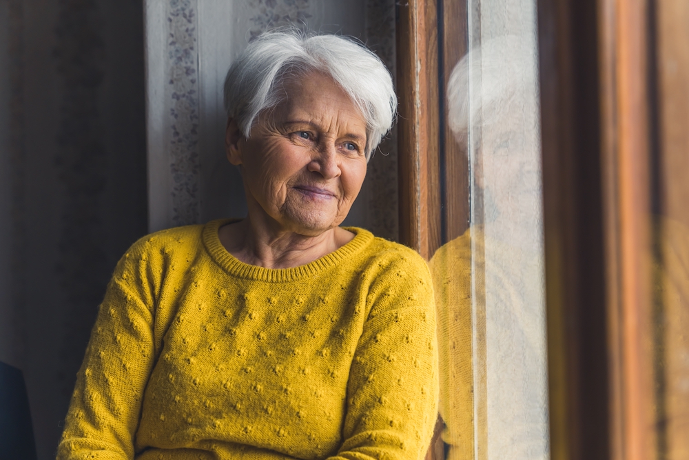 Une femme âgée dans un chandail jaune regarde par sa fenêtre